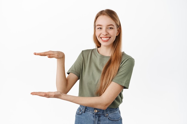 Free photo young smiling female model showing an item holding something empty space between hands looking pleased recommending product standing over white background