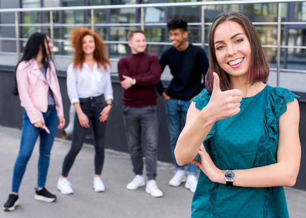 Young smiling female showing thumb up gesture looking at camera while her friends standing blurred background