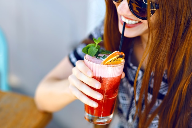 Young smiling girl drinking tasty sweet cocktail , amazing relaxing day, tasty lemonade, elegant dress and sunglasses, outdoor terrace.