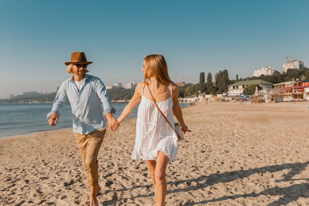 Young smiling happy man in hat and blond woman running together on beach on summer vacation traveling