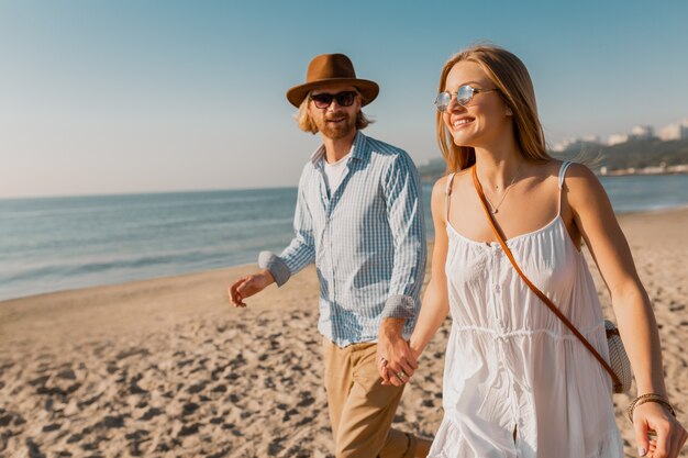 Young smiling happy man in hat and blond woman running together on beach on summer vacation traveling