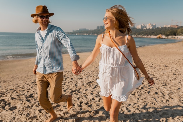 Young smiling happy man in hat and blond woman in white dress running together on beach on summer vacation traveling