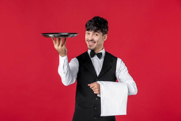 Free photo young smiling male waiter in a uniform with butterfly on neck and raising tray holding towel on red background