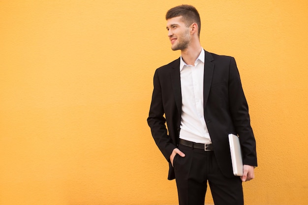 Young smiling man in classic black suit and white shirt holding laptop in hand while dreamily looking aside over orange background