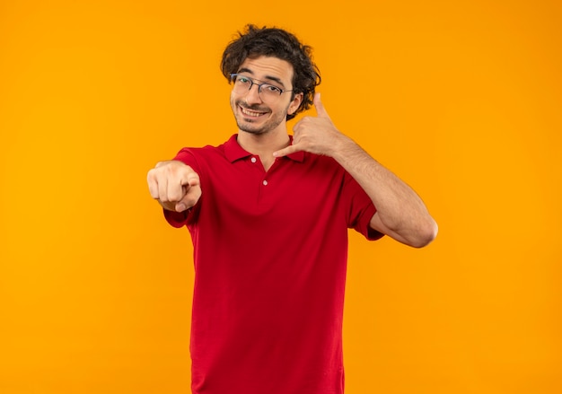 Free photo young smiling man in red shirt with optical glasses gestures call me hand sign and points isolated on orange wall