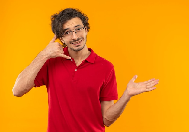 Free photo young smiling man in red shirt with optical glasses gestures call me hand sign and points at side isolated on orange wall