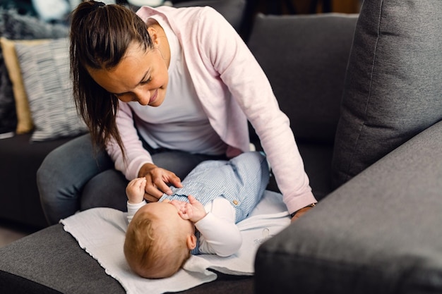 Young smiling mother taking care of her baby boy who is lying on the sofa