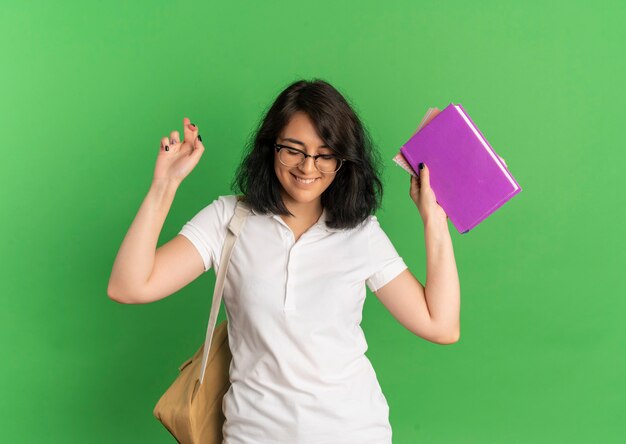 Young smiling pretty caucasian schoolgirl wearing glasses and back bag crosses fingers holding books looking down on green  with copy space