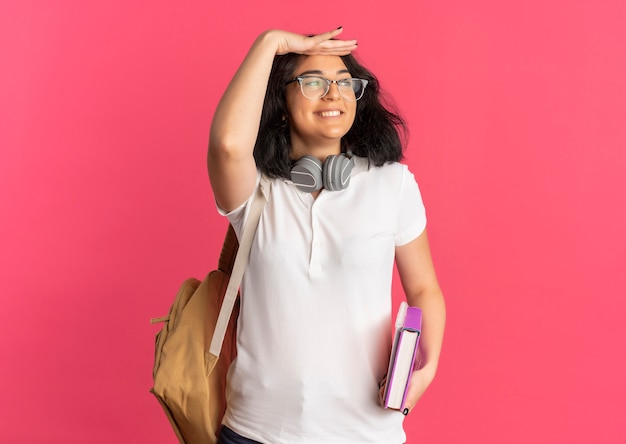 Free Photo young smiling pretty caucasian schoolgirl wearing glasses back bag and headphones on neck keeping palm at her forehead trying to see something holding book and notebook