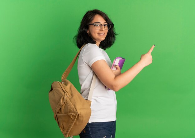 Young smiling pretty caucasian schoolgirl wearing glasses and back bag stands sideways points at side on green  with copy space