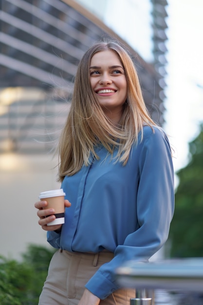 Young smiling professional woman having a coffee break during her full working day. She holds a paper cup outdoors near the business building while relaxing and enjoying her beverage.