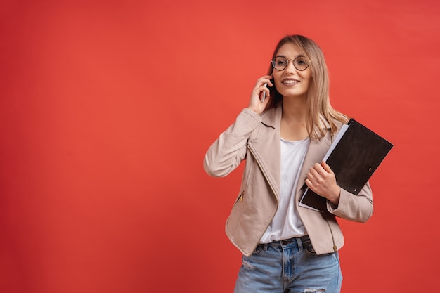 Young smiling student or intern in eyeglasses talking on the phone while standing with a folder