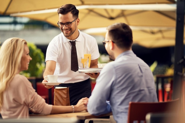 Young smiling waiter brining coffee to a couple in an outdoor cafe