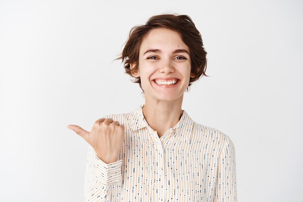 Young smiling woman employee looking happy and proud, pointing finger left company, standing against white wall