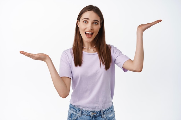 Young smiling woman, female customer weighing in hands, raising open palms sideways as if holding two items, products to choose, white background