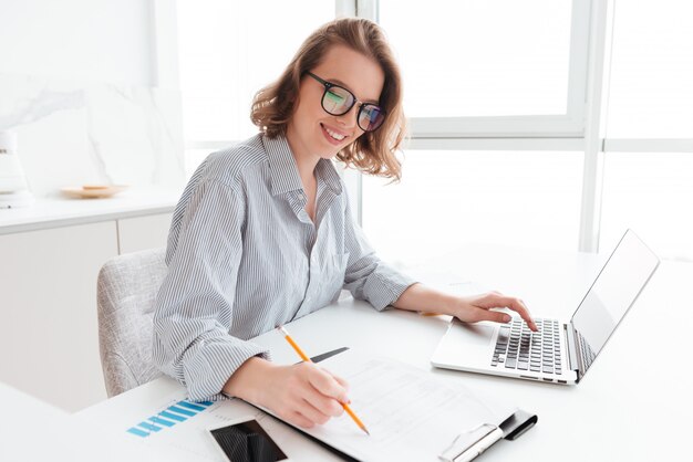 Young smiling woman in glasses and striped shirt working with documents and computer while siting at table in light kitchen