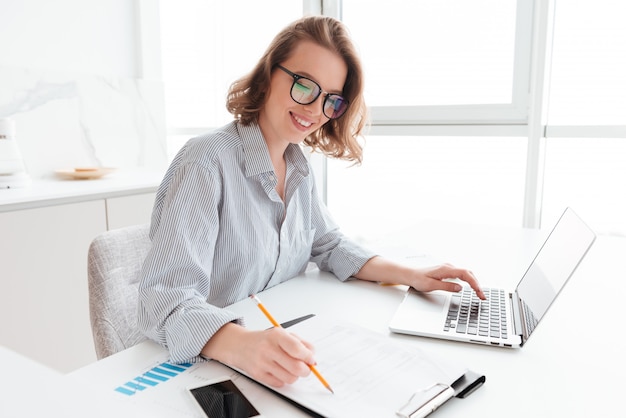 Young smiling woman in glasses and striped shirt working with documents and computer while siting at table in light kitchen