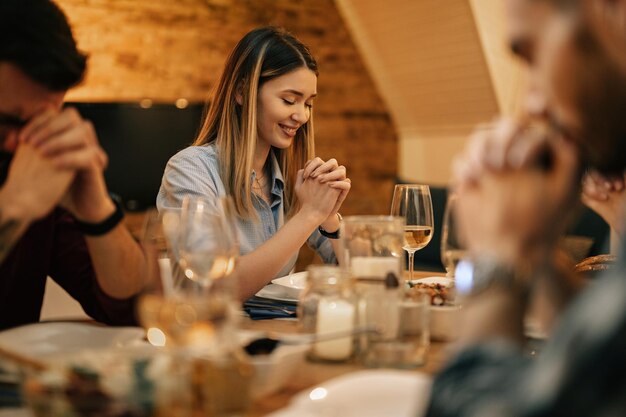 Young smiling woman and her friends sitting at dining table and praying before a meal