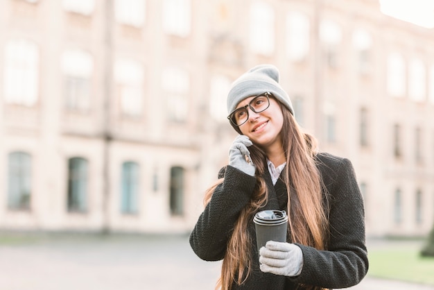 Young smiling woman with cup talking on smartphone on street