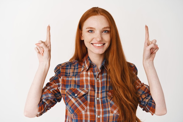 Young smiling woman with long ginger hair and blue eyes, pointing fingers up with happy face