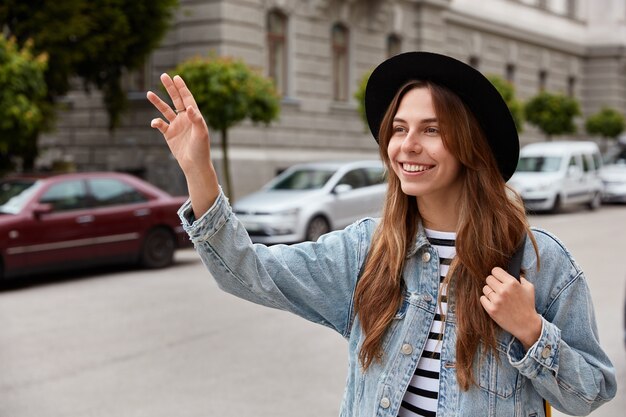 Young smiling young European woman walks outdoor, waves hand as notices friend into distance