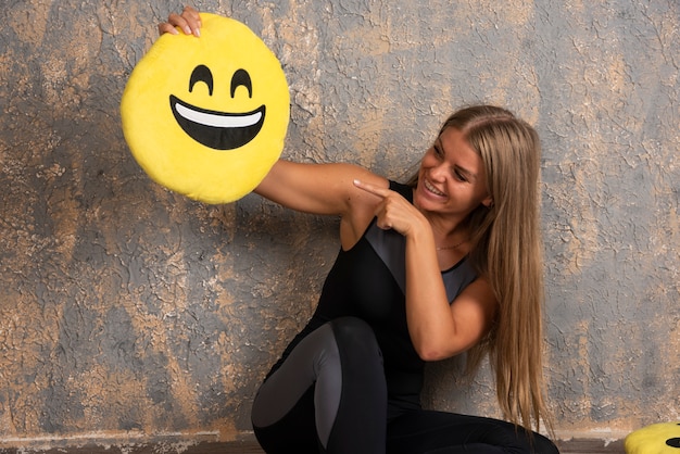 Free Photo young sportive girl in sport outfits holding a smiling emoji pillow and pointing at it.