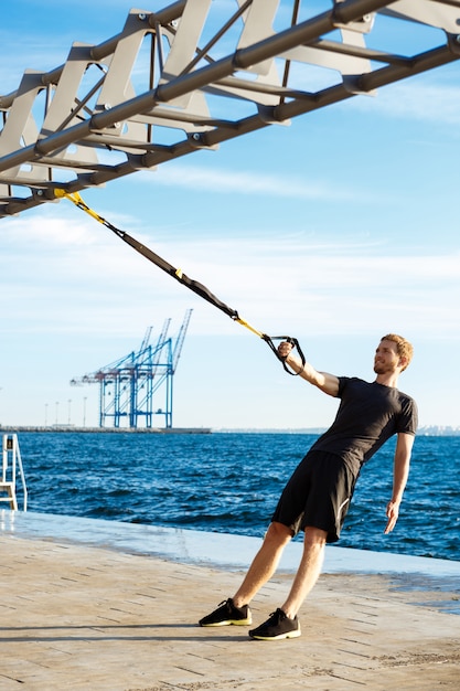 Free photo young sportive man training with trx near sea in the morning.