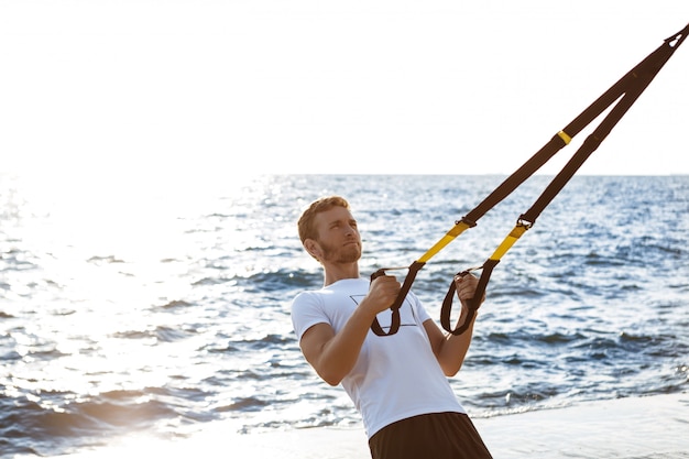 Young sportive man training with trx near sea in the morning.