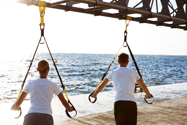 Free photo young sportive people training with trx near sea in the morning.