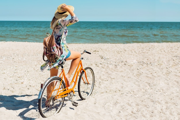 Free photo young sportive woman in stylish white crop top and denim shorts standing on the beach with orange bicycle