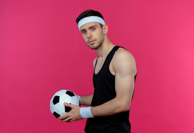 Young sporty man in headband holding soccer ball looking at camera with serious expression standing over pink background