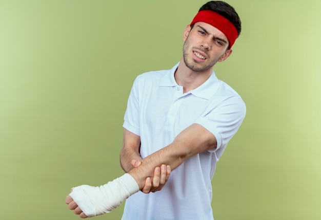 Free photo young sporty man in headband touching his bandaged hand feeling pain over green