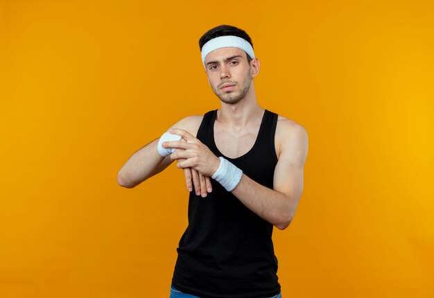 Young sporty man in headband touching his wrist stretching his hands standing over orange background