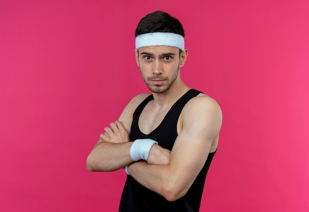 Free photo young sporty man in headband with confident serious expression with crossed arms on chest over pink