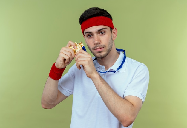 Young sporty man in headband with gold medal around neck showing his medal looking confident over green