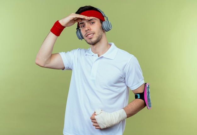 Free photo young sporty man in headband with headphones and smartphone arm band looking far away with hand over head standing over green background