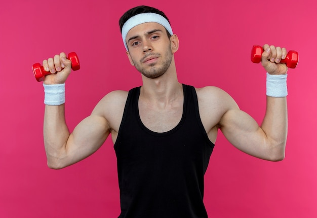 Young sporty man in headband working out with dumbbells strained and confident over pink