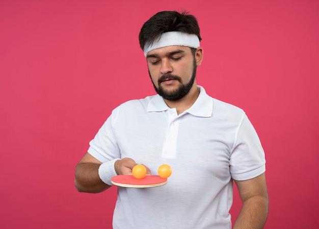 Young sporty man wearing headband and wristband holding and looking at ping pong racket with balls