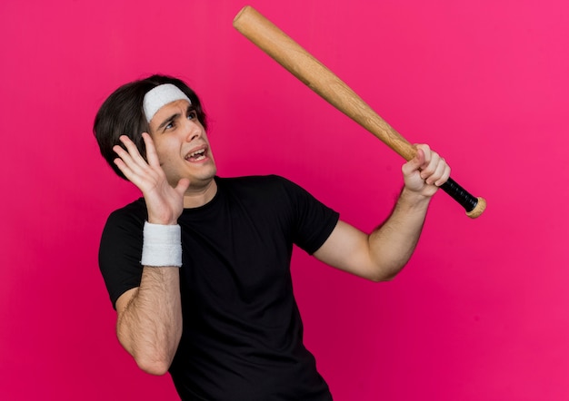 Young sporty man wearing sportswear and headband holding baseball bat looking at something scared making defense gesture with hand 