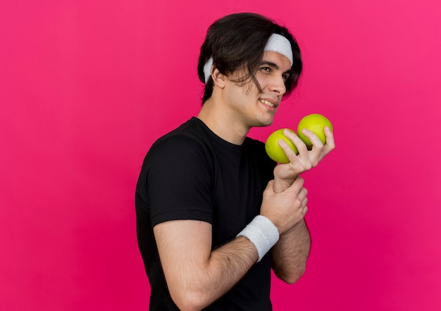 Young sporty man wearing sportswear and headband holding green apples looking aside with smile on face 