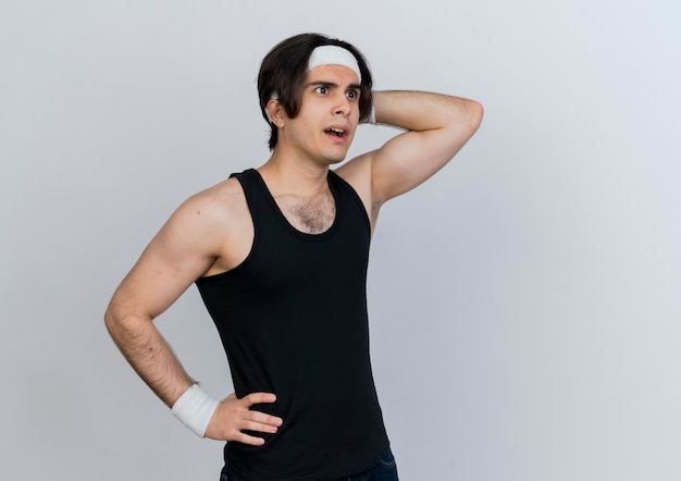 Free photo young sporty man wearing sportswear and headband looking aside puzzled with hand on his head standing over white wall