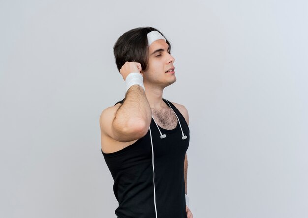 Young sporty man wearing sportswear and headband looking aside tired standing over white wall