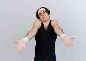 Free photo young sporty man wearing sportswear and headband looking at front confused with arms out standing over white wall