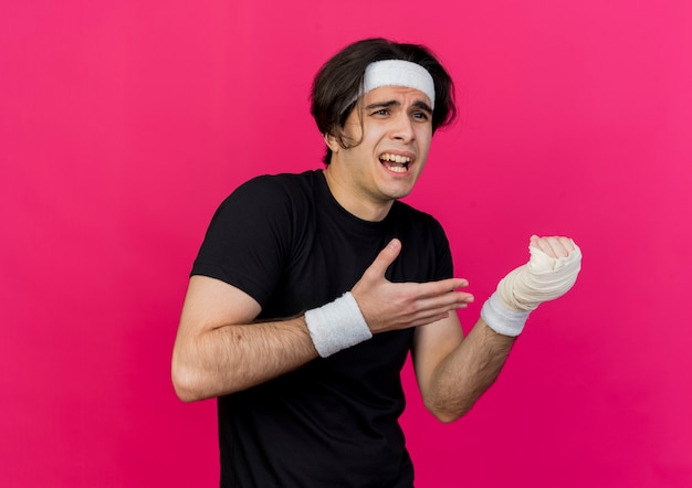 Young sporty man wearing sportswear and headband pointing with arm at his bandaged wrist feeling pain 