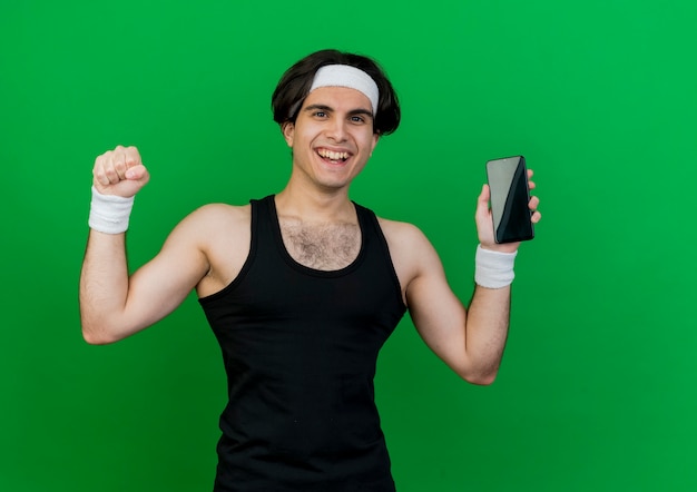 Young sporty man wearing sportswear and headband showing smartphone clenching fist happy and excited 