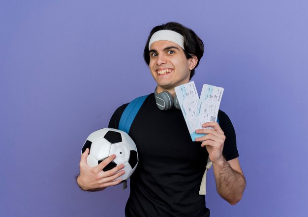 Young sporty man wearing sportswear and headband with backpack holding soccer ball and air tickets smiling with happy face 