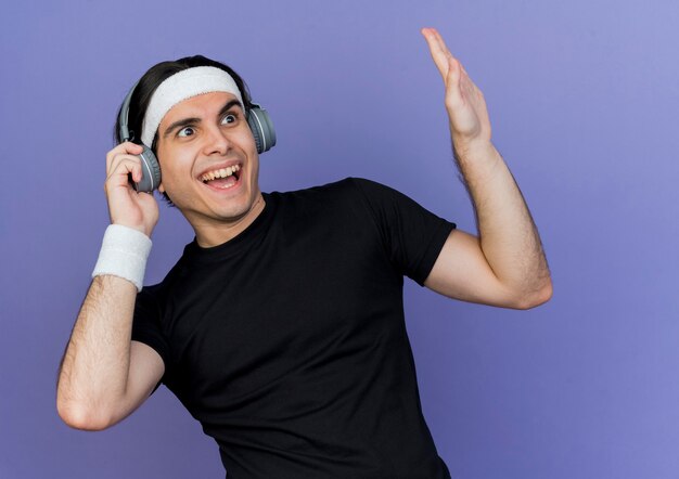 Young sporty man wearing sportswear and headband with headphones looking aside happy and excited gesturing with hand 