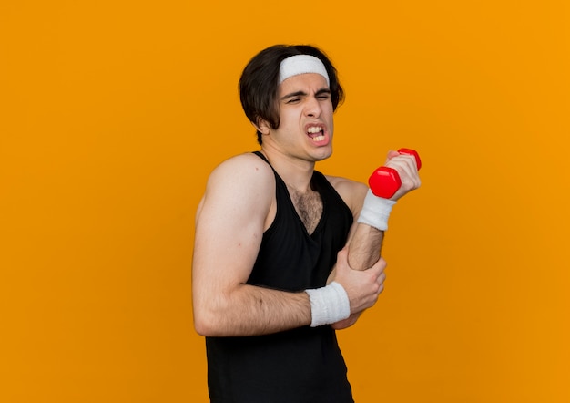 Young sporty man wearing sportswear and headband working out with dumbbell looking confused and displeased standing over orange wall