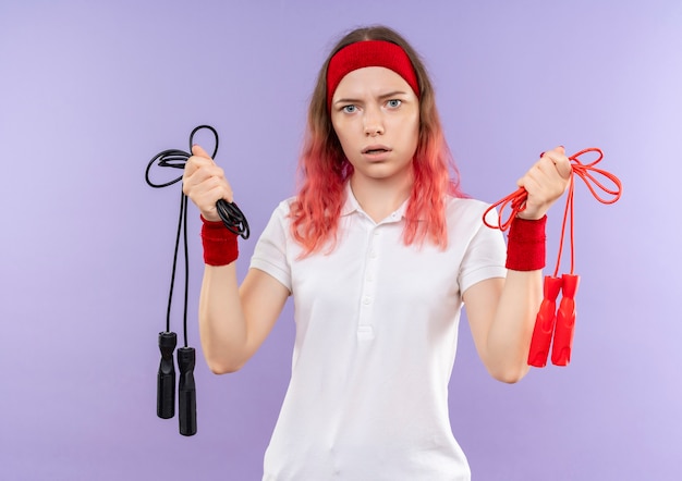 Free photo young sporty woman in headband holding skipping ropes confused standing over purple wall