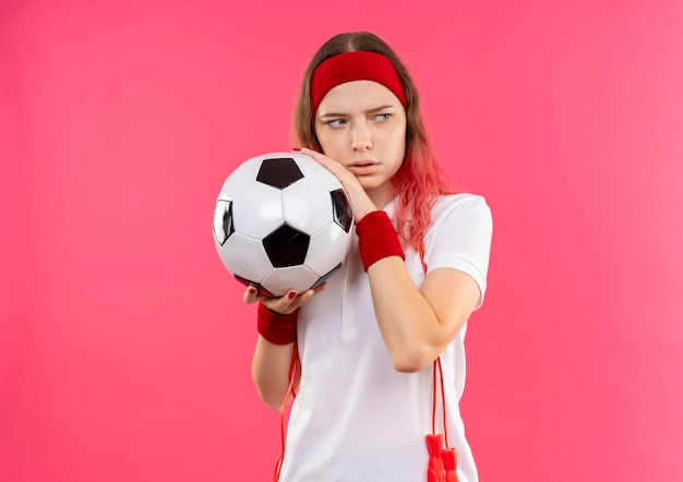 Free Photo young sporty woman in headband holding soccer ball looking aside with fear expression standing over pink wall